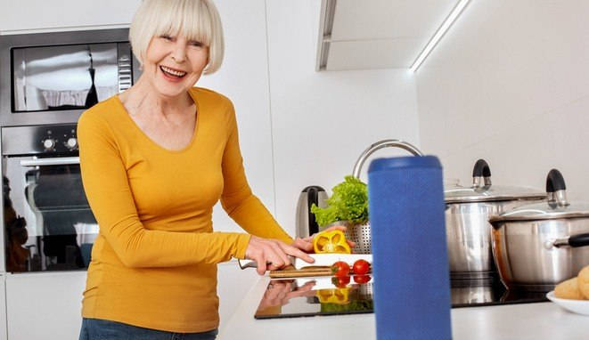 Retirement. Senior woman standing at kitchen cooking dinner asking to search recipe with wireless smart speaker smiling cheerful