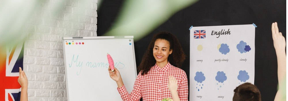 Educación, Enseñanza de la Lengua Inglesa, audio lingual method; una profesora joven señala a la pizarra con palabras simples en inglés, los niños tienen las manos levantadas