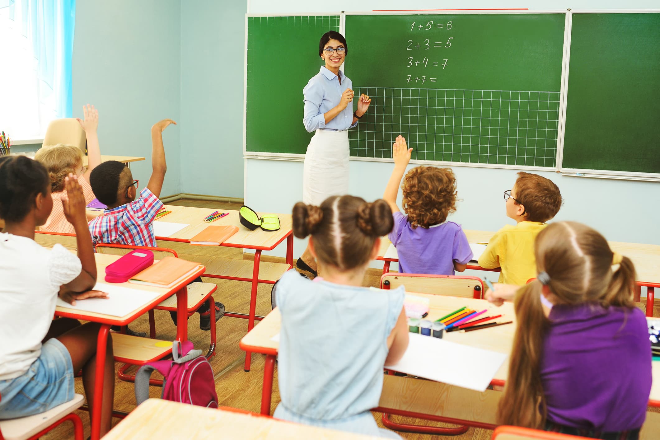 Los niños en edad preescolar levantan las manos para responder a la pregunta del maestro sobre el fondo de la pizarra y el aula en la escuela primaria