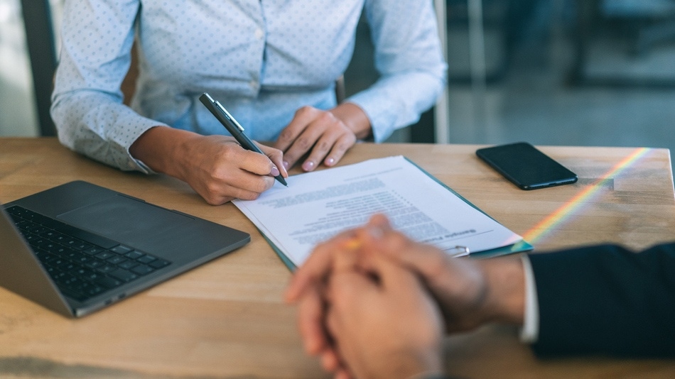 Mujer firmando un contrato frente a un hombre con las manos cogidas sobre la mesa