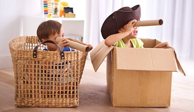 Niños jugando en casa. Uno metido en la cesta de la compra y otro en una caja de cartón 