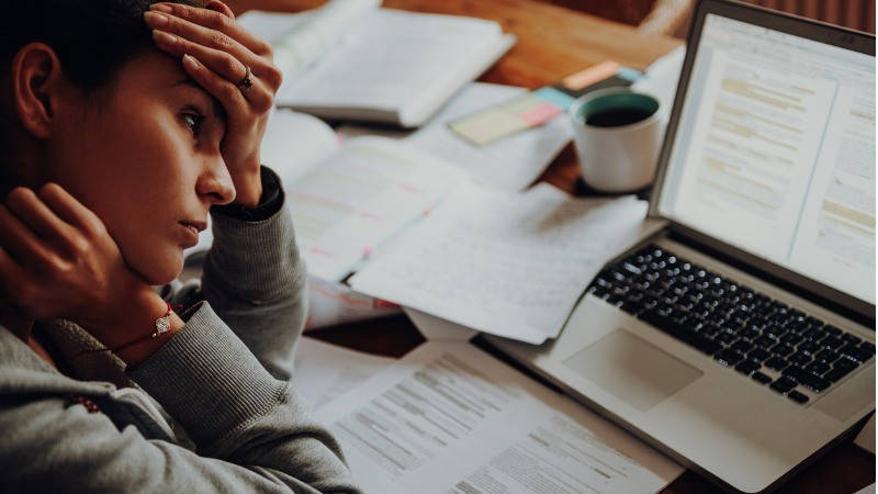 mujer estudiando rodeada por papeles y el ordenador, con dolor de cabeza por ansiedad