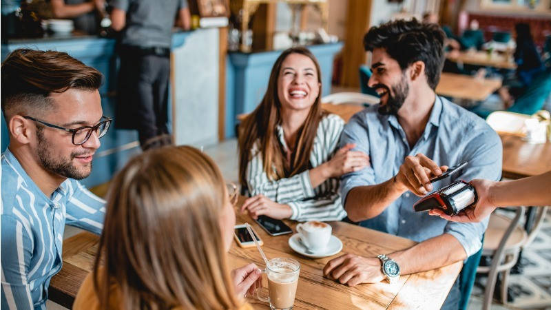Medios de pago, contactless; un grupo de amigos riéndose en un café mientras uno paga con su móvil