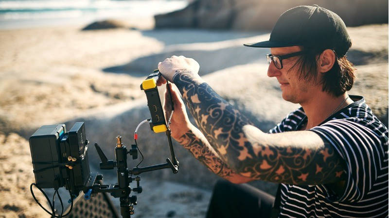 Transmedia storytelling, narrativa transmedia; rodaje, un chico con gorra, gafas y tatuajes graba con un videocámara en la playa