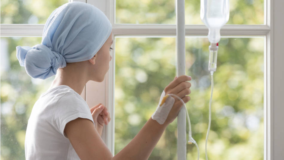 leucemia en niños, una joven con pañuelo en la cabeza y bolsa IV mira por la ventana